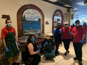 Masked restaurant workers pose in front of a table with food packaged to go